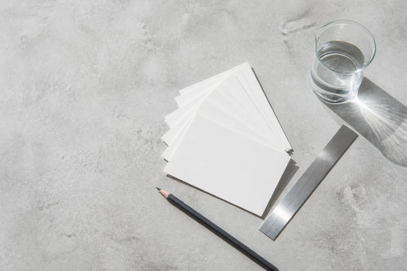 Overhead view of blank white index cards fanned in an arc on a pale concrete surface, beside a graphite pencil, a thin steel rule and a plain glass beaker of water casting a bright caustic shadow.